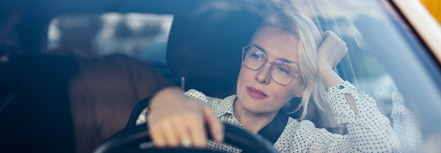 A woman sits in a car with one hand on the wheel and an elbow on the side window with her hand holding her head up.