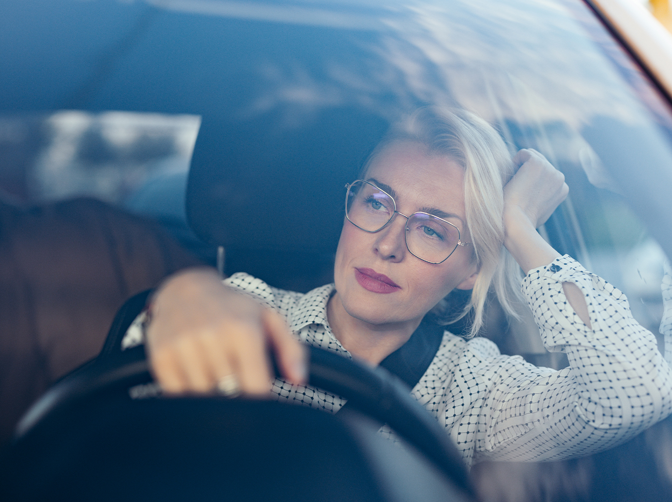 A woman sits in a car with one hand on the wheel and an elbow on the side window with her hand holding her head up.