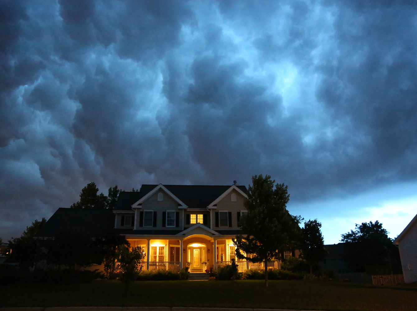 A lit suburban home at night beneath a dramatic dark storm sky.
