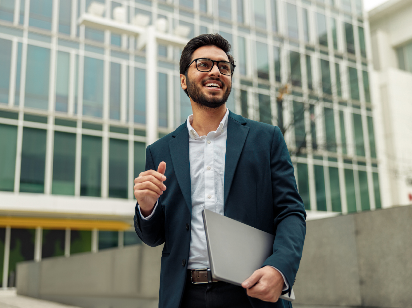 A smiling young professional in a blazer holds a laptop outside a modern glass office building.