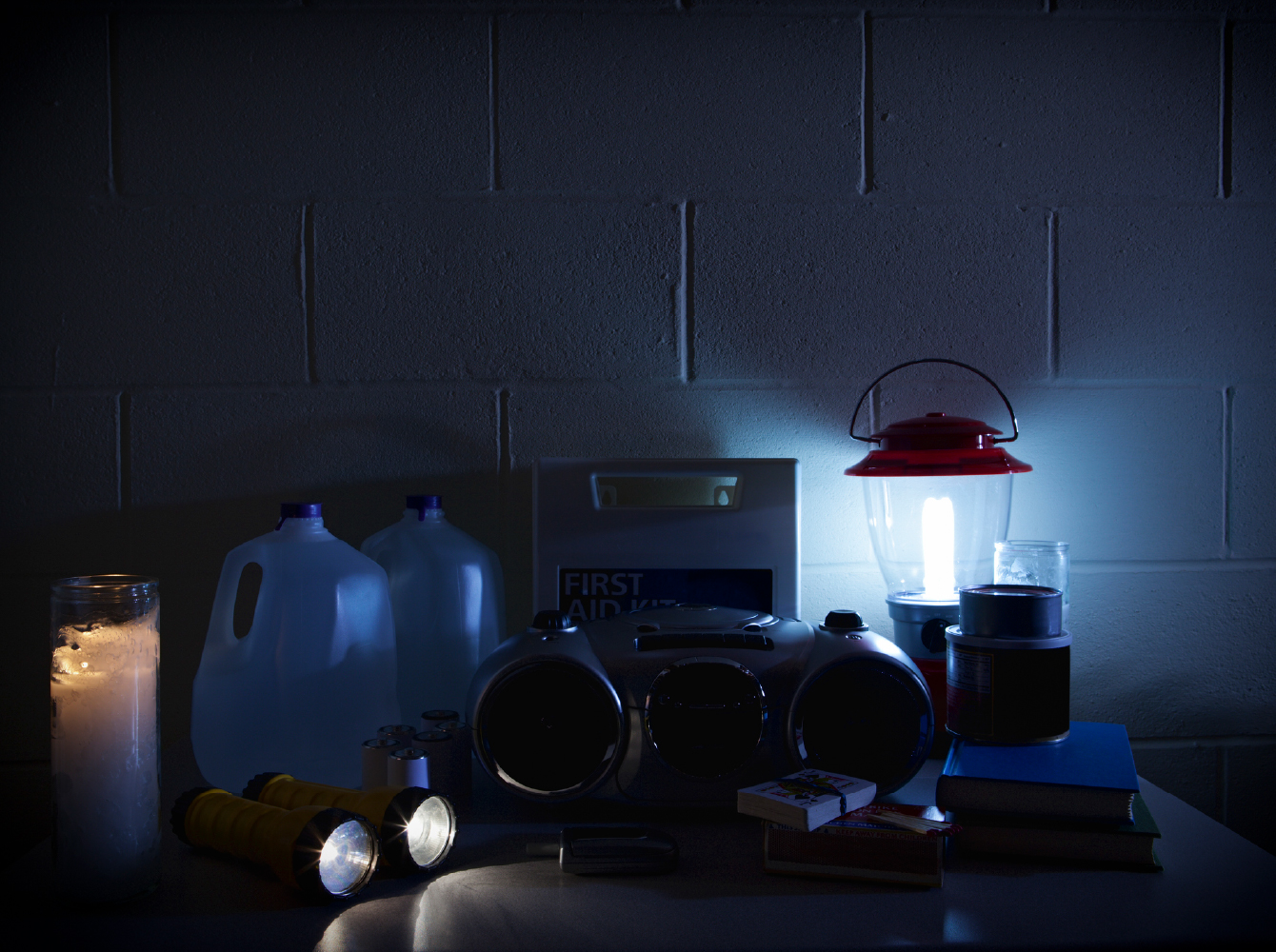Power outage supplies including a lantern, candle, flashlights, water jugs and a first aid kit arranged in a dark room.