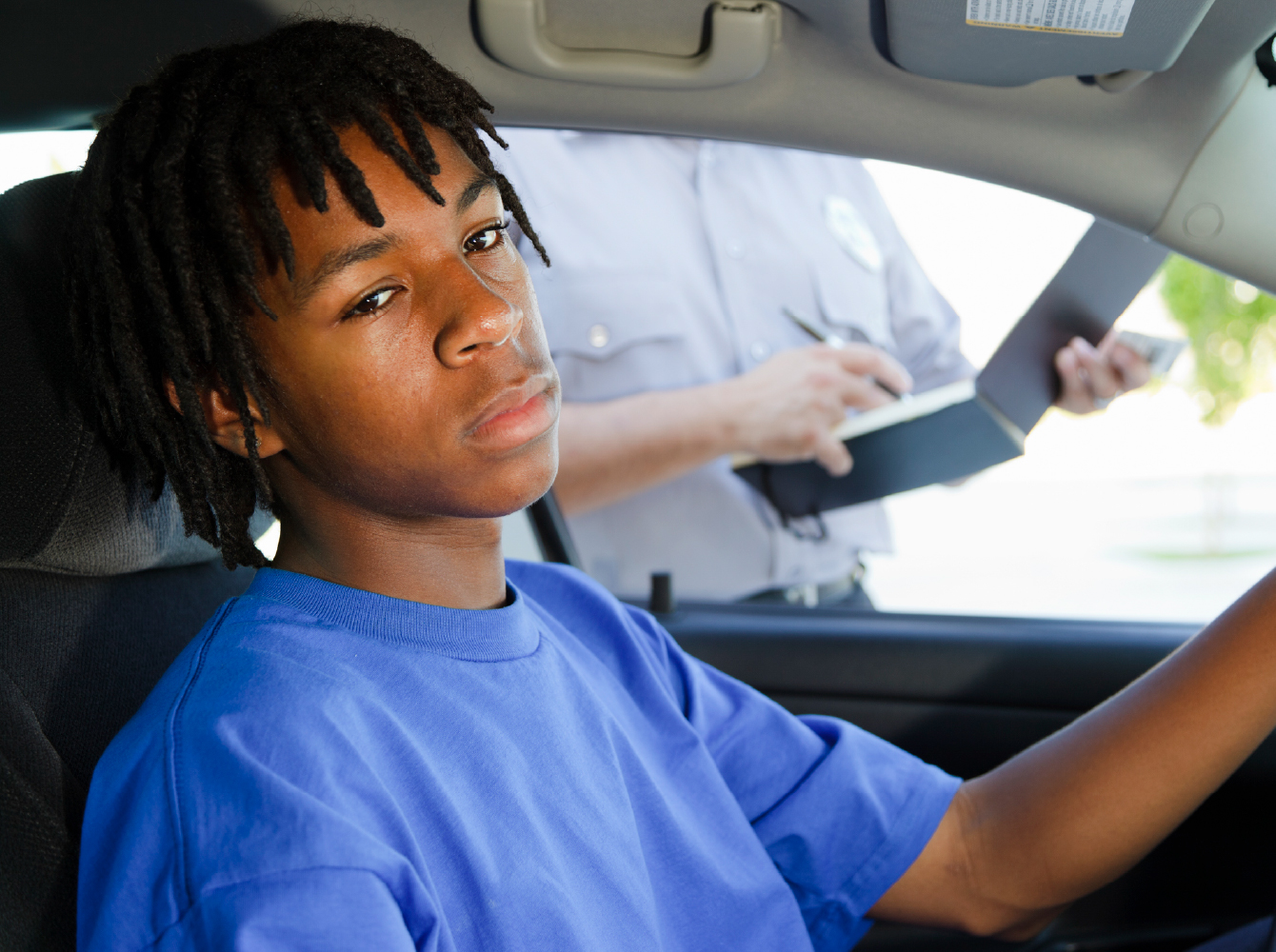 A young driver sits in a car looking apprehensive while an officer writes a ticket outside the window.
