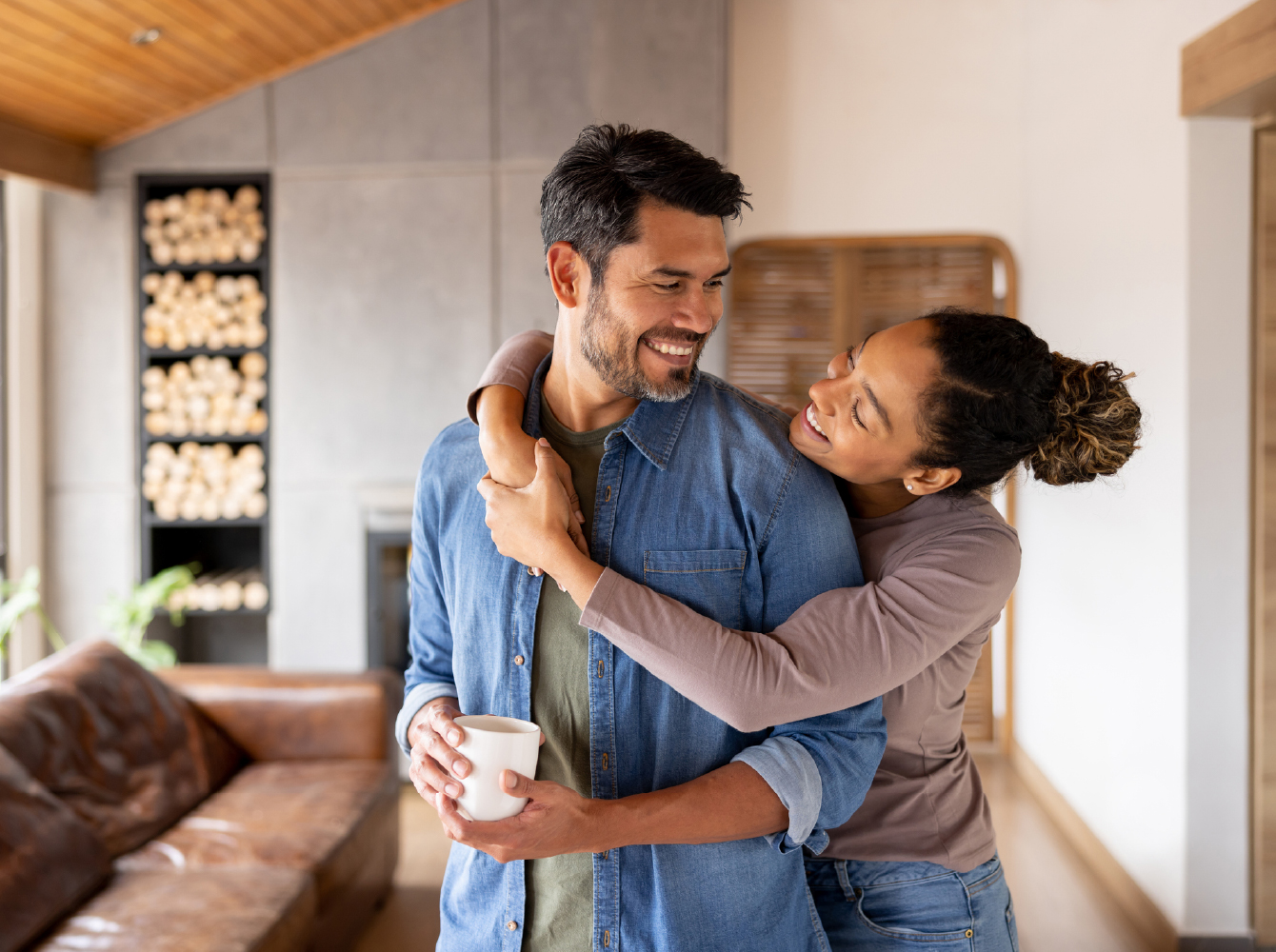 Two people smiling and embracing in a cozy living room, one holding a coffee mug.