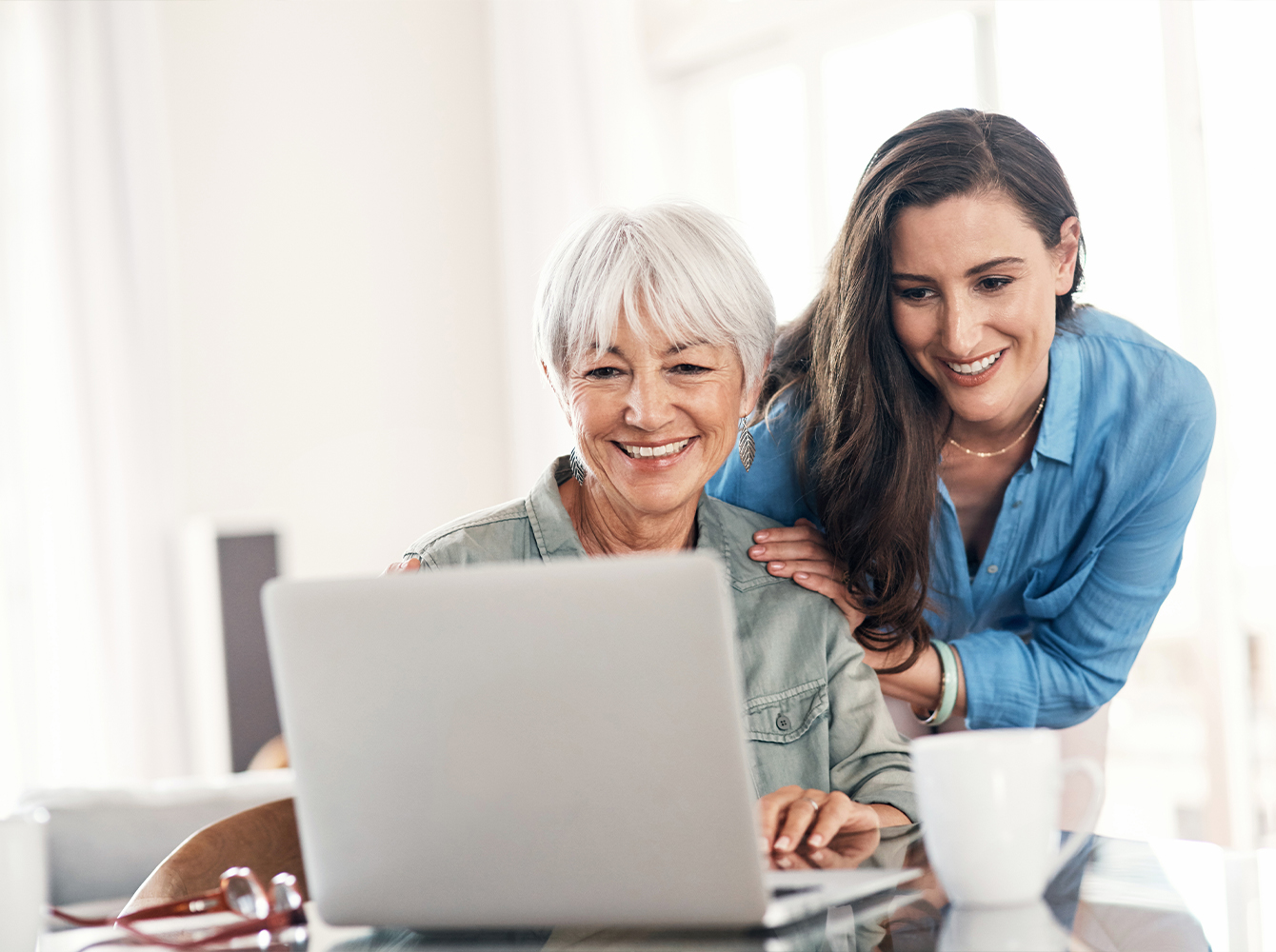 A younger woman stands behind an older woman helping her use a laptop.