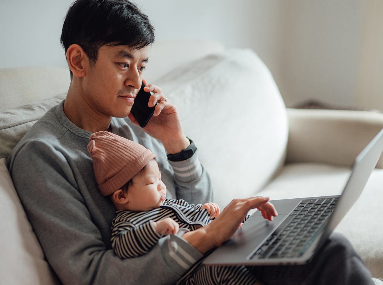 A man with a baby in his lap holds a cellphone to his ear while working on a laptop.