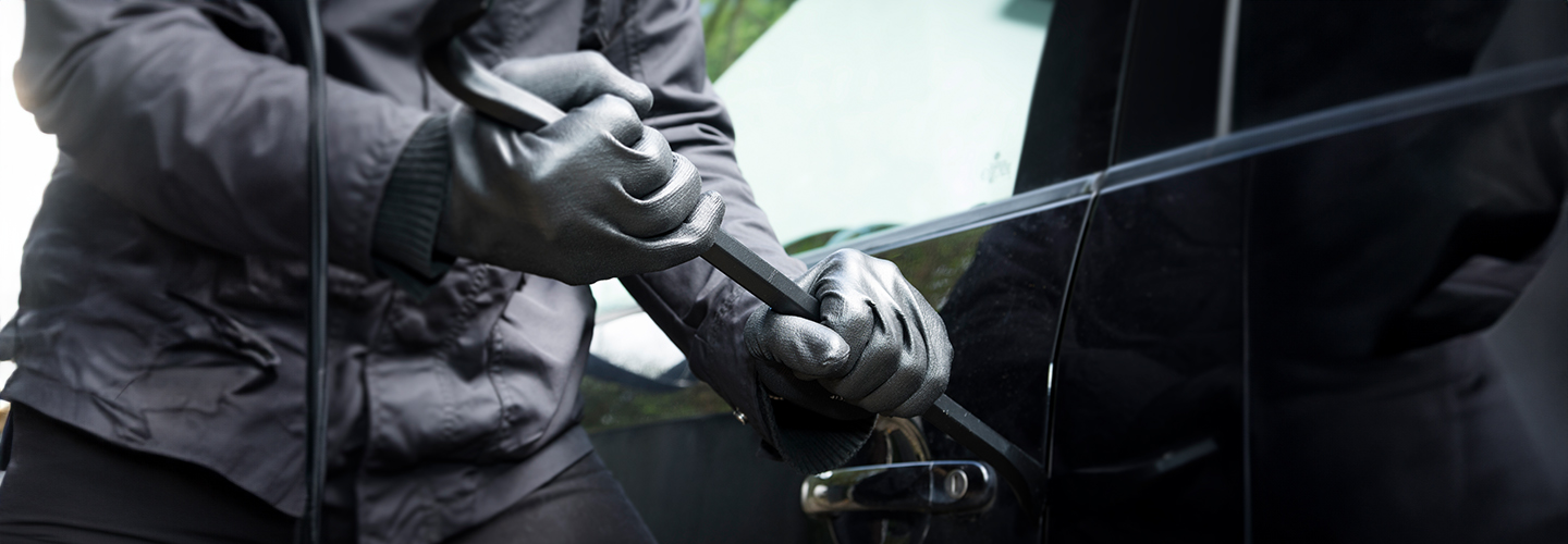 A person in all black with gloves on is using a crow bar to try to break into a car.