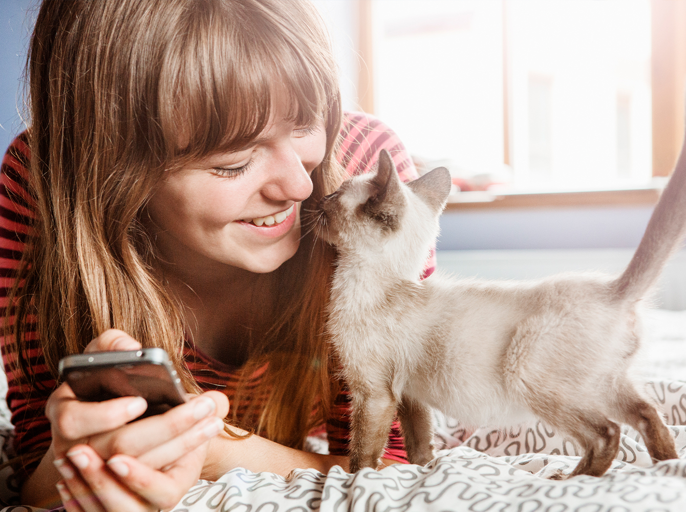 A young woman is laying on a bed with a phone in her hand while a kitten comes up to sniff her.