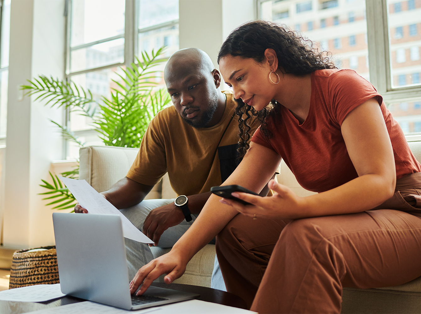 A couple is sitting down in front of a laptop while looking at a phone and reviewing documents.