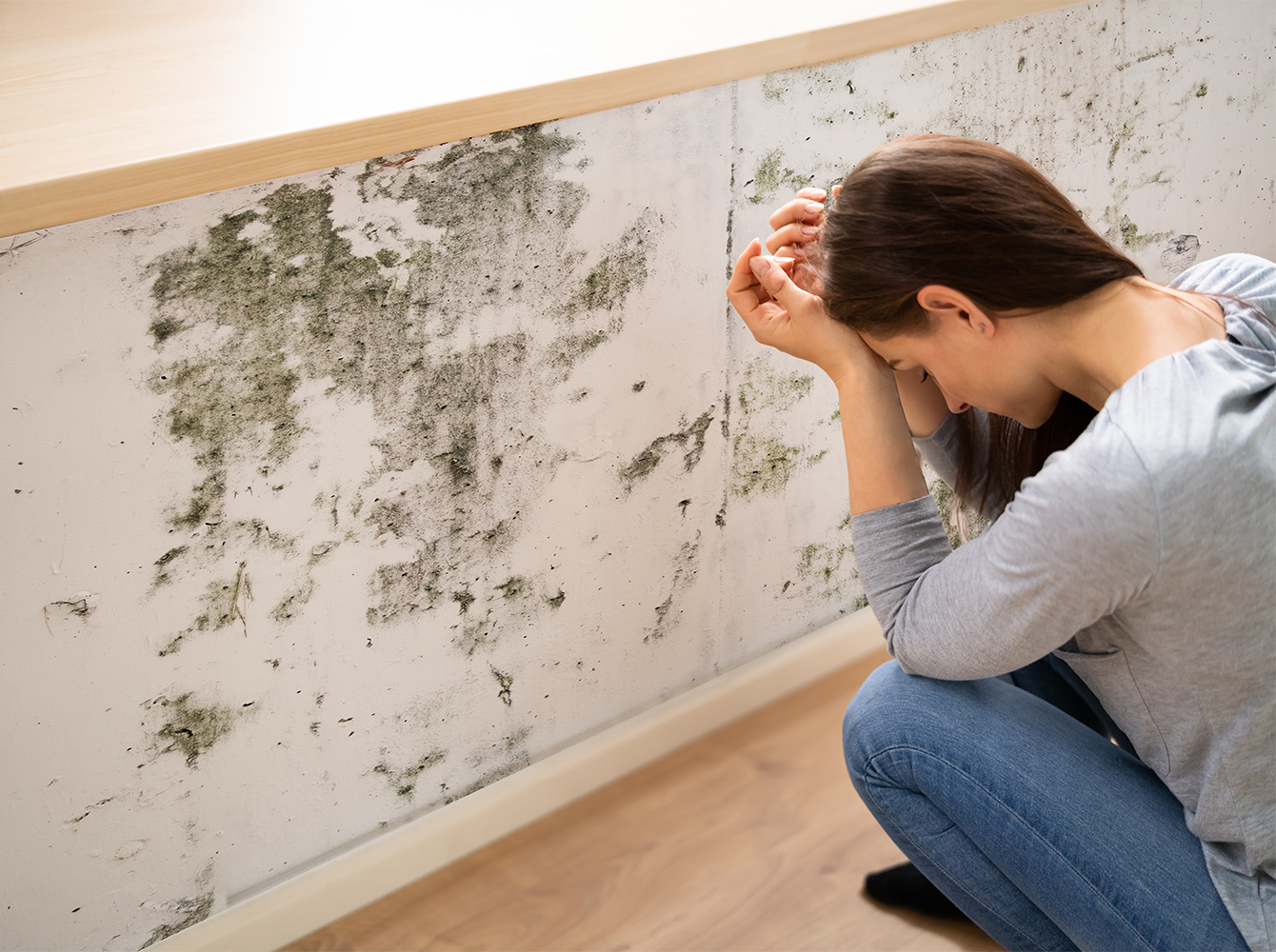 A woman is crouching in front of a moldy wall with her head in her hands.