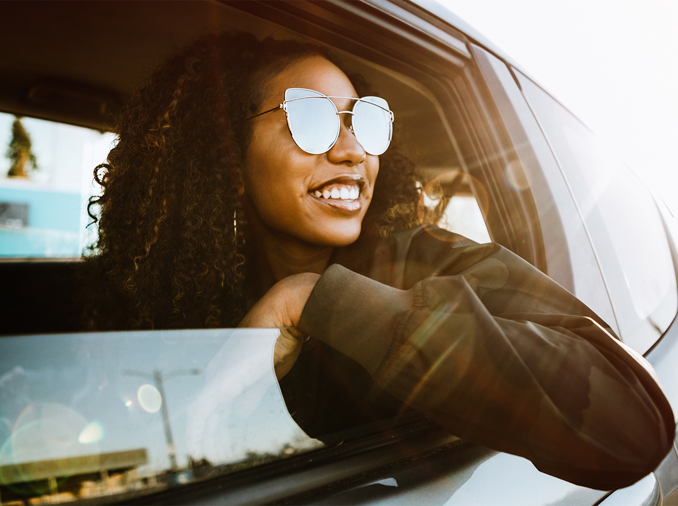 A smiling woman with sunglasses on is looking out the window of a car.