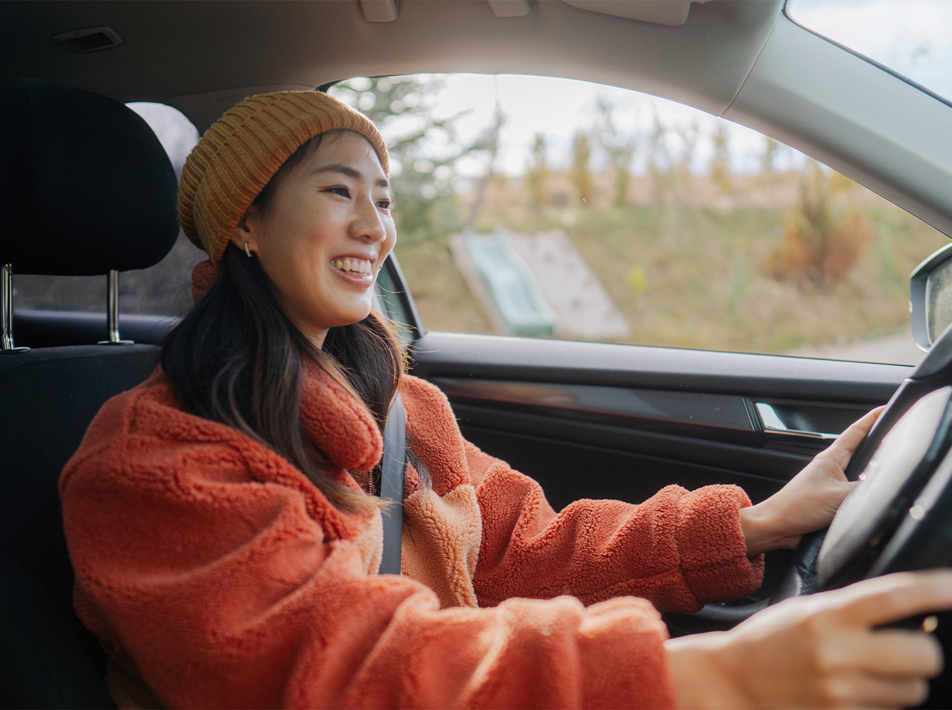 A smiling woman in a sweatshirt and beanie is driving a car.
