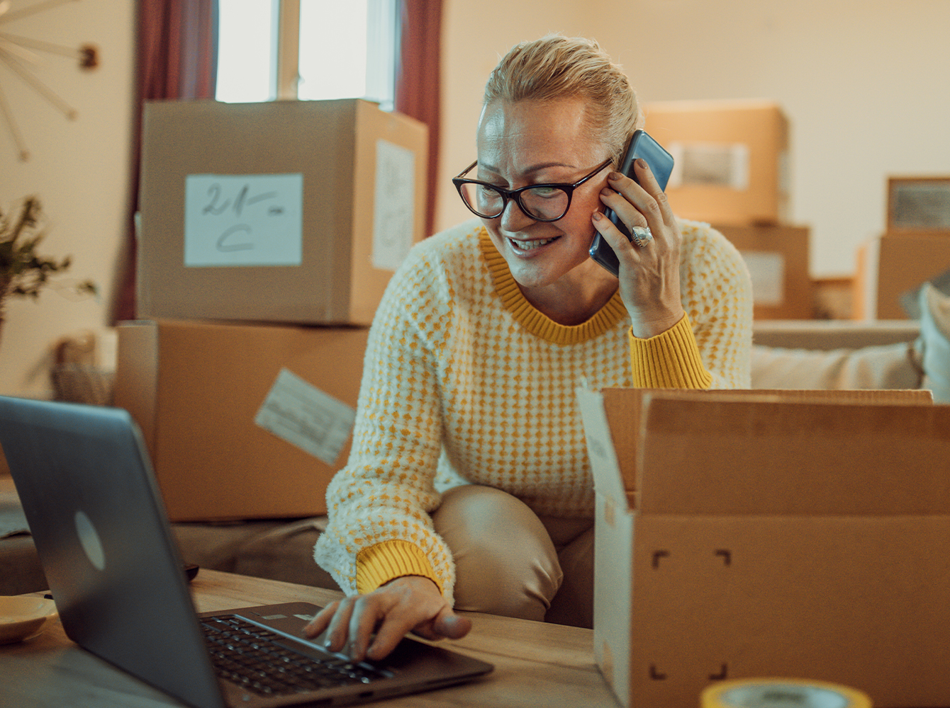 A woman is on the phone and sitting in a room filled with moving boxes.
