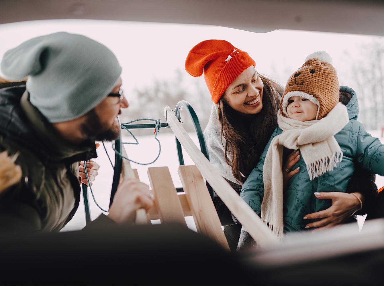 A family dressed in winter apparel is behind their car. The man is unloading a sled, while the woman is holding their toddler.
