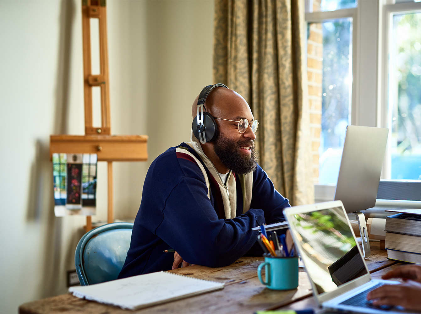A smiling man with headphones on is sitting at a cluttered table, looking at a laptop.