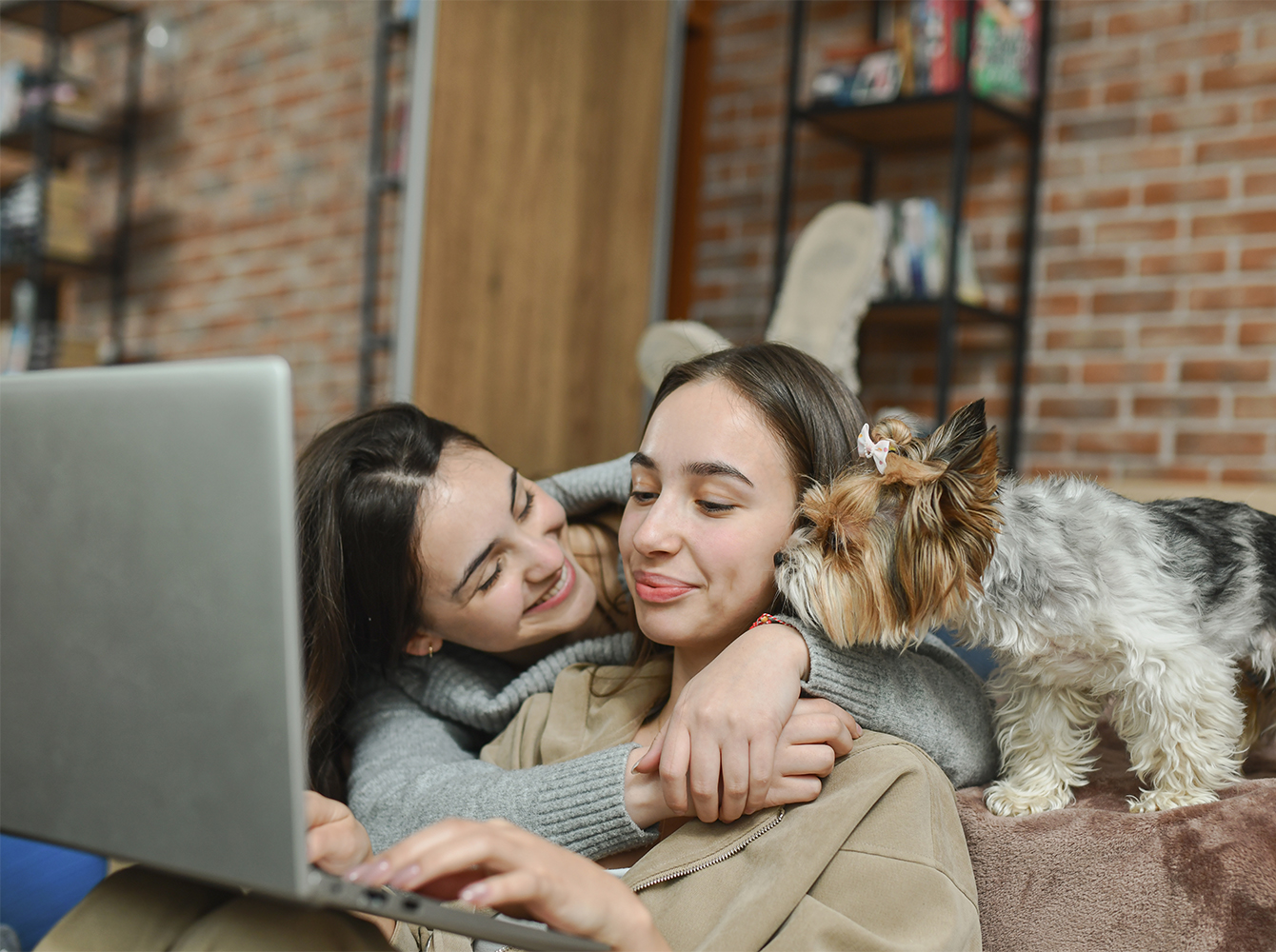 A mother hugs her daughter while they look at a laptop and a dog looks on.