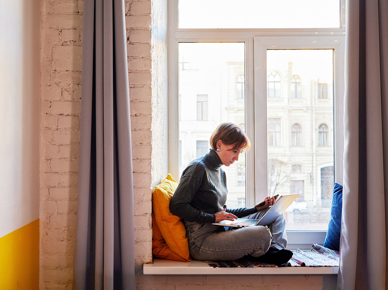 A person is sitting on a window seat looking at a laptop.