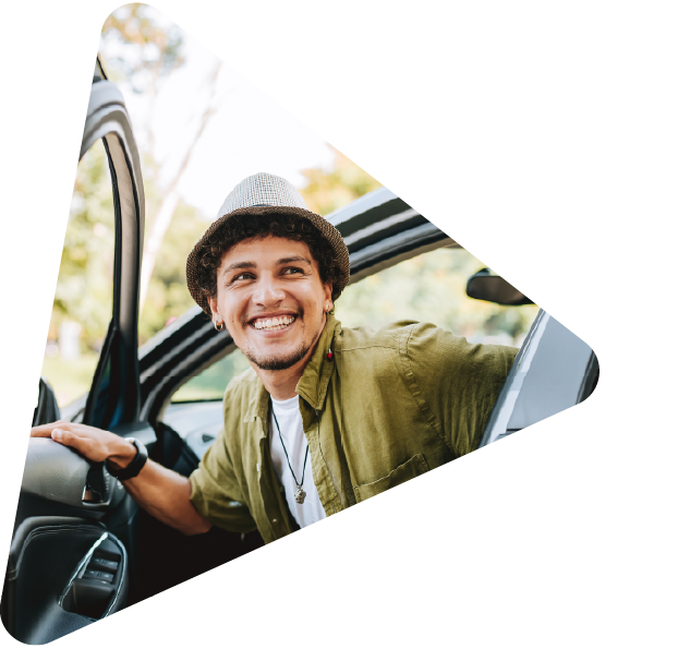 A smiling young man in a hat sits in the driver's seat of a car with the door open.
