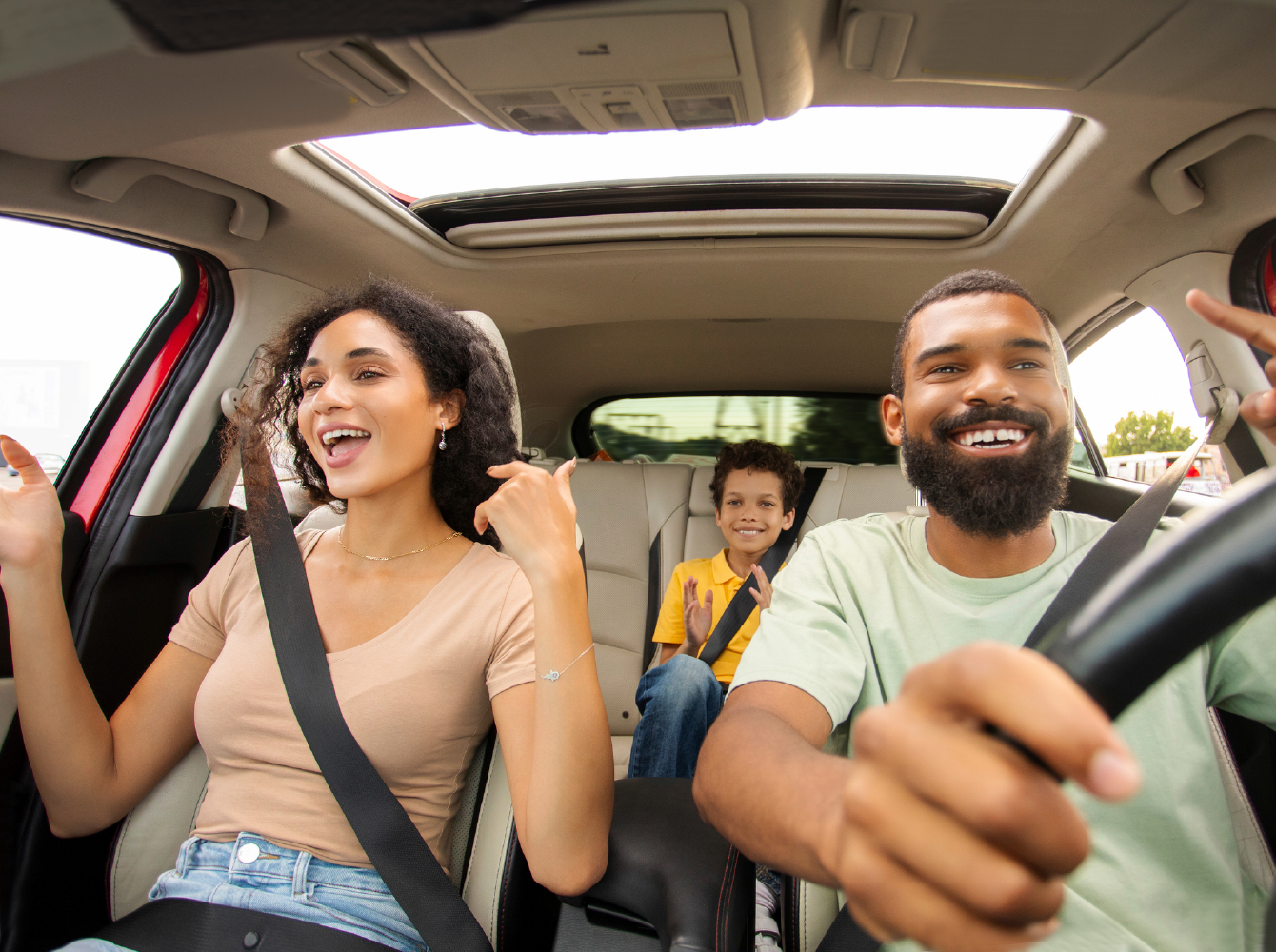 A laughing family of three enjoys a road trip, with two adults in the front seat and a smiling child in the back.