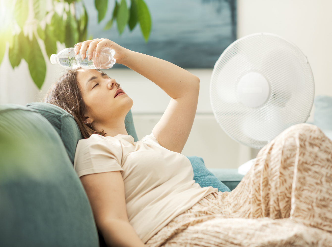 A woman cooling off on a sofa by pressing a cold water bottle to her forehead in front of a fan.