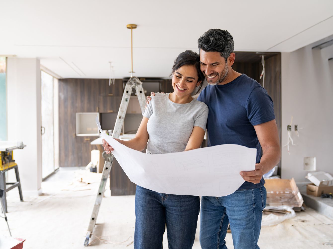 Two people smiling and reviewing blueprints inside a home mid-renovation with a ladder and tools visible.