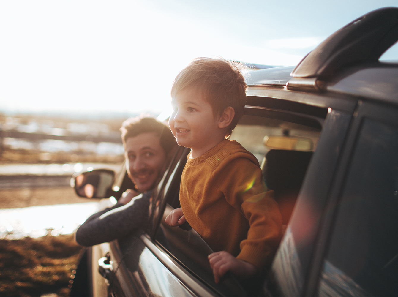 A father and son are sticking their heads out of a car window on a sunny day.