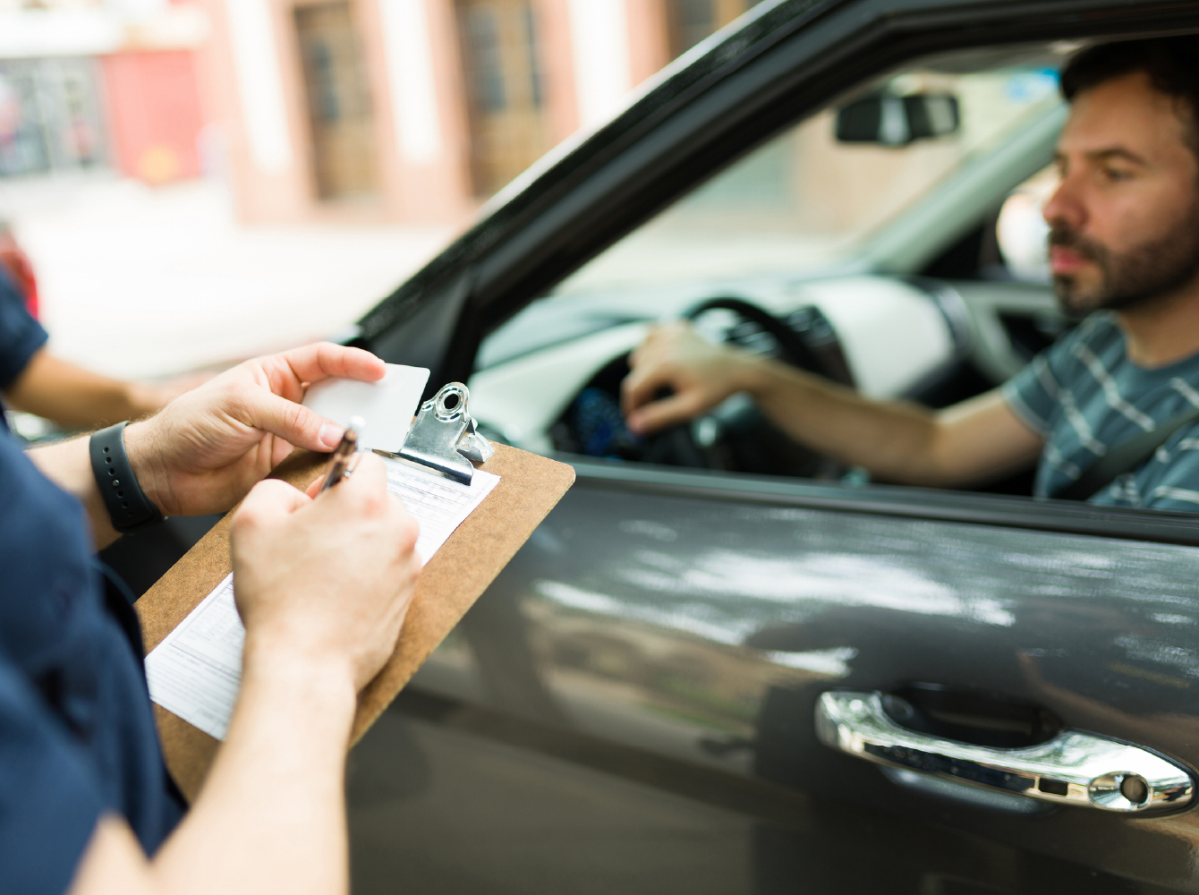 An officer writing on a clipboard while accepting a driver's license from a man seated in a pulled-over vehicle.