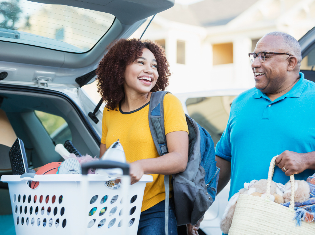 A smiling young woman with a backpack and laundry basket unloads a car with an older man, suggesting a student moving away from home.