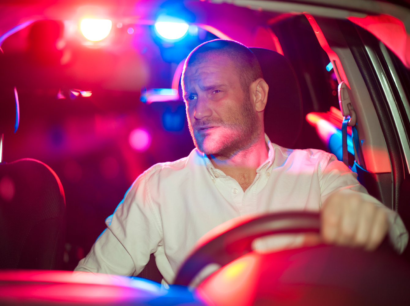 A tense man gripping a steering wheel at night, lit by red and blue police lights behind his vehicle.