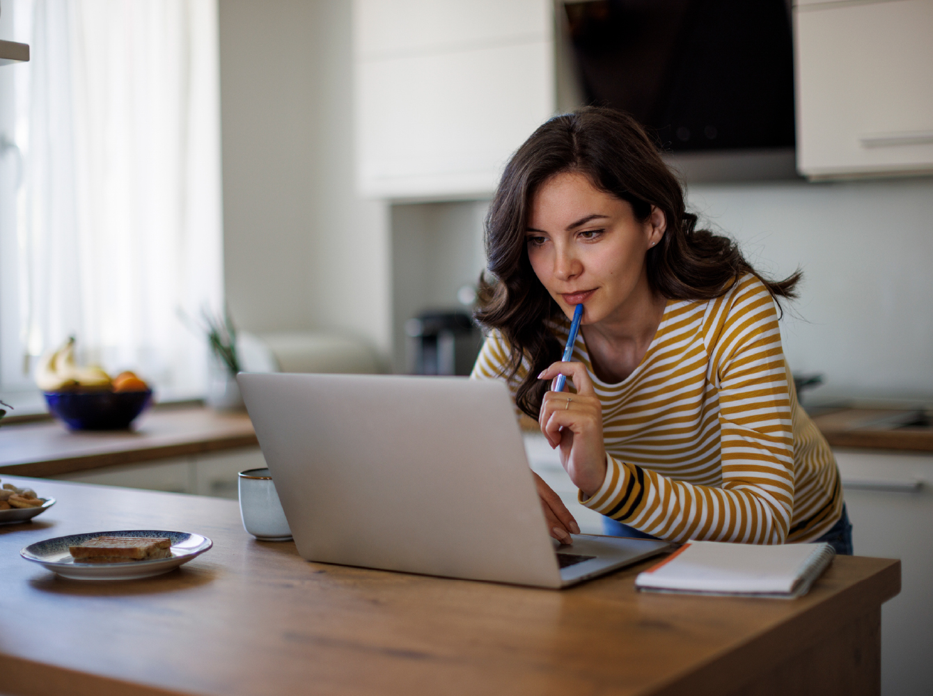A woman pensively reviews information on a laptop at a kitchen table with a notepad nearby.