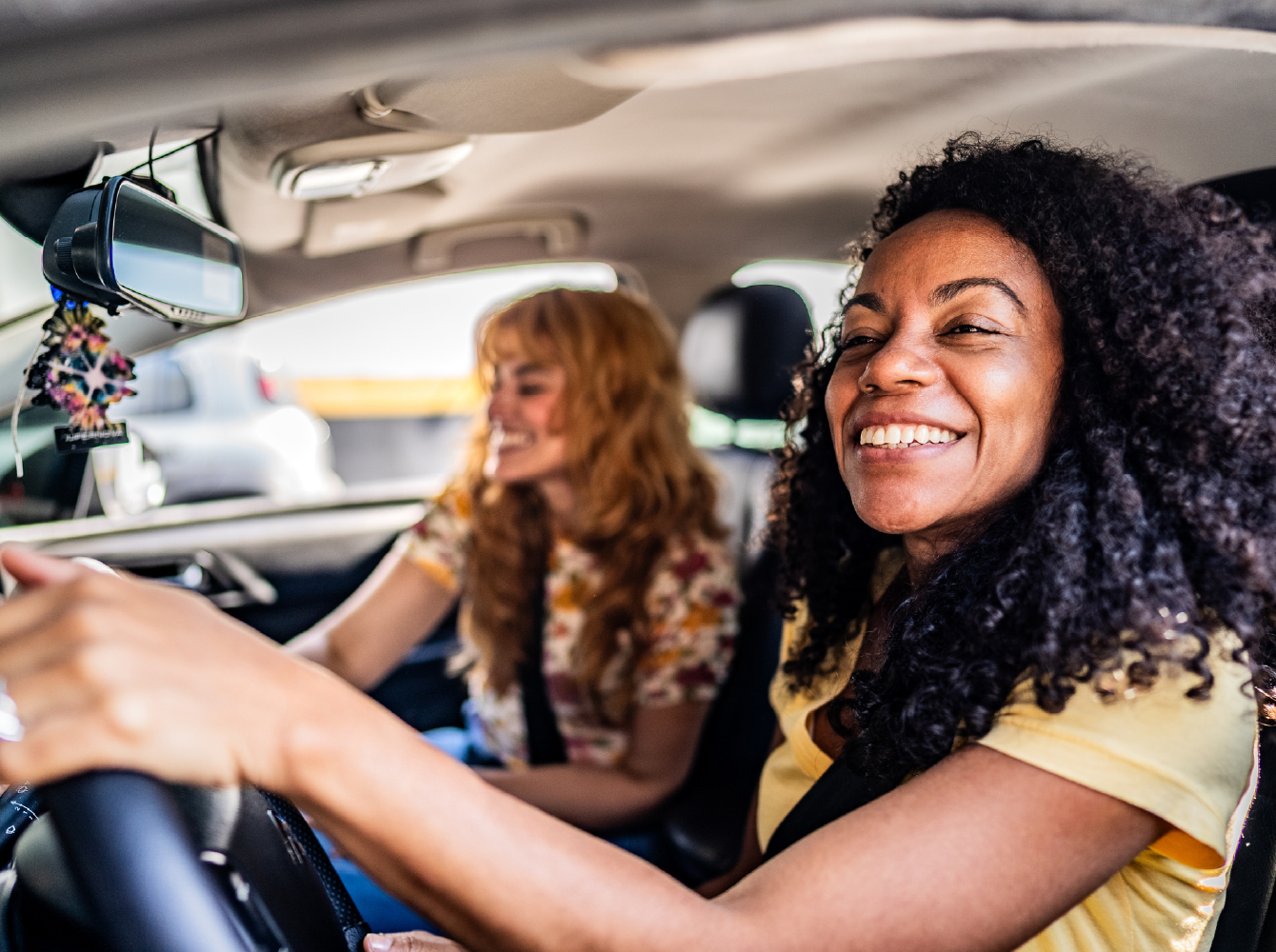 A smiling woman drives a car with a laughing passenger beside her.