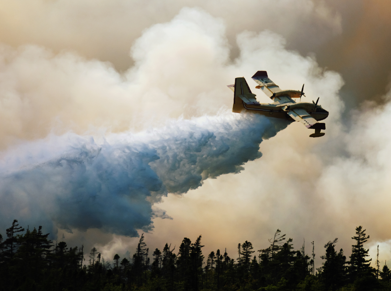 A water bomber aircraft drops retardant over a forest amid thick wildfire smoke.