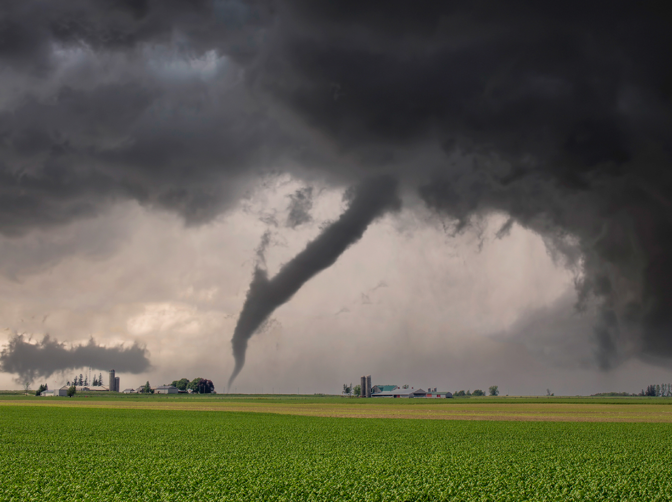 A large tornado touches down near farm buildings across an open green field under dark storm clouds.