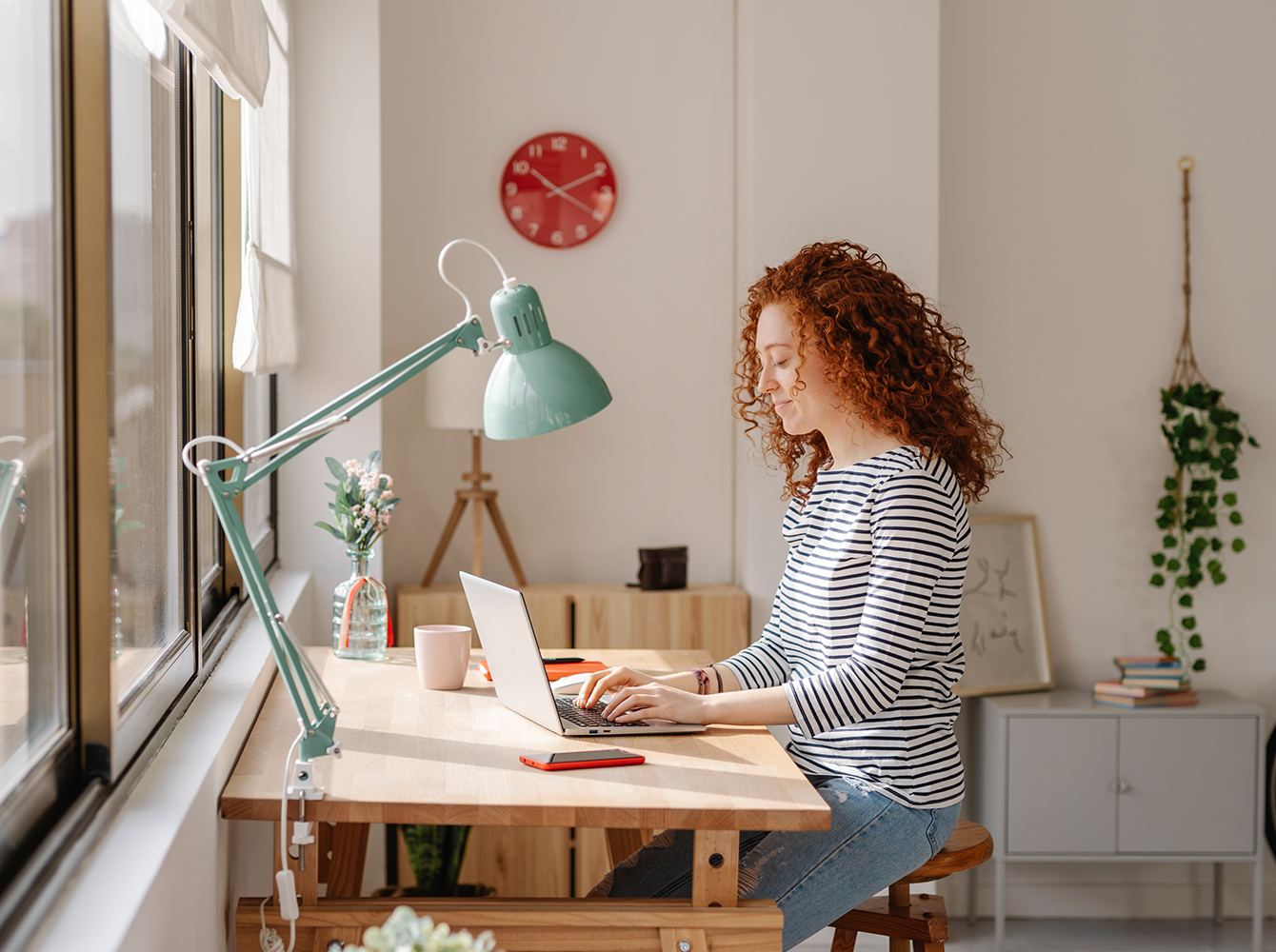 A woman is sitting at a desk by a window working on a laptop.