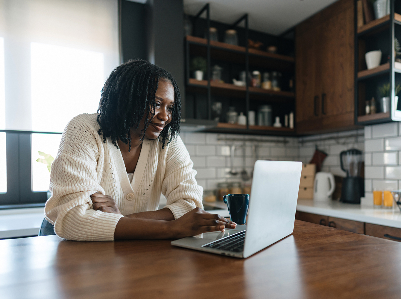 A smiling woman is sitting in her kitchen, looking at her laptop on the table in front of her.