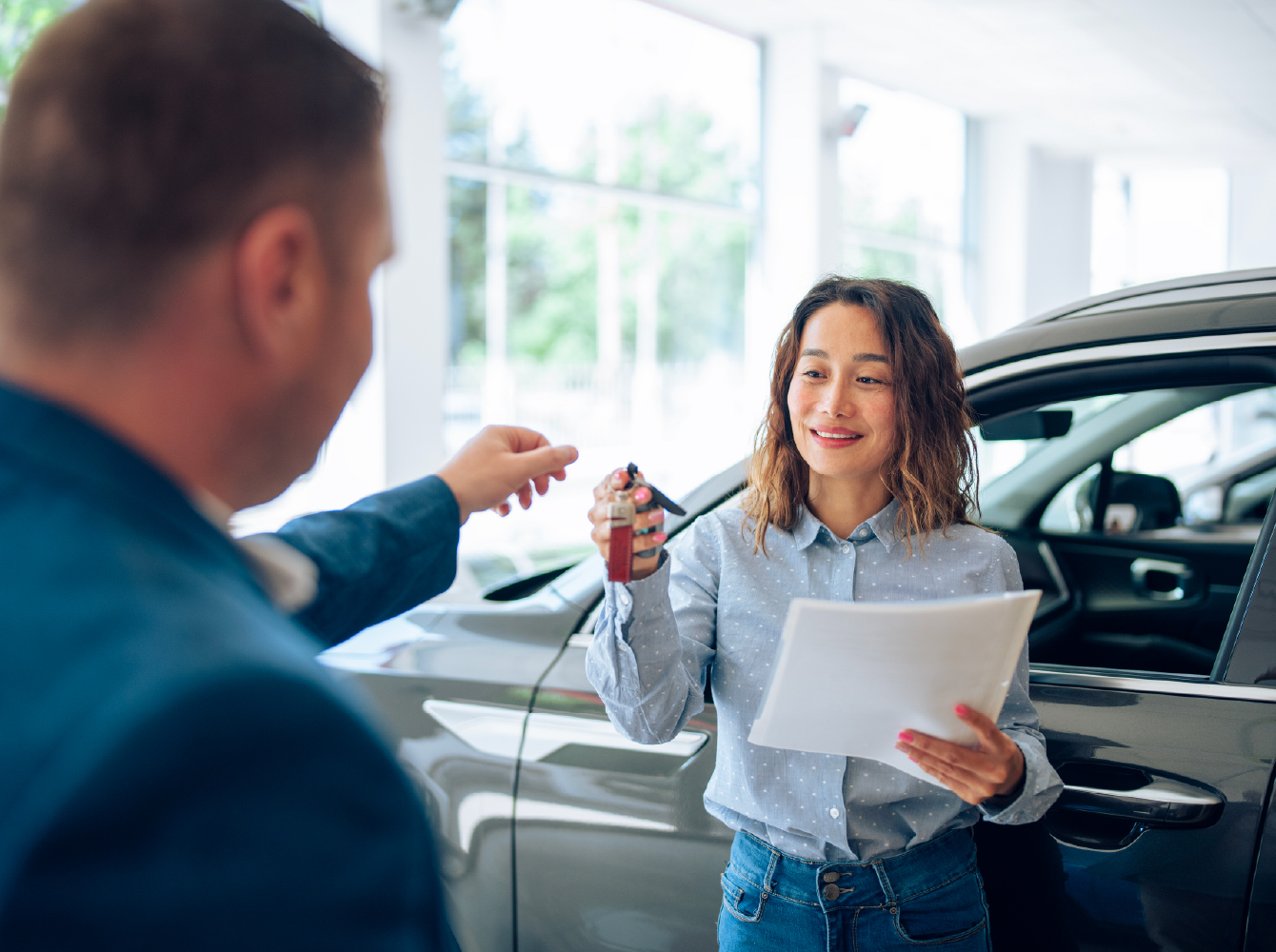 A smiling woman holding documents receives car keys from a person at a dealership, with a dark sedan in the background.