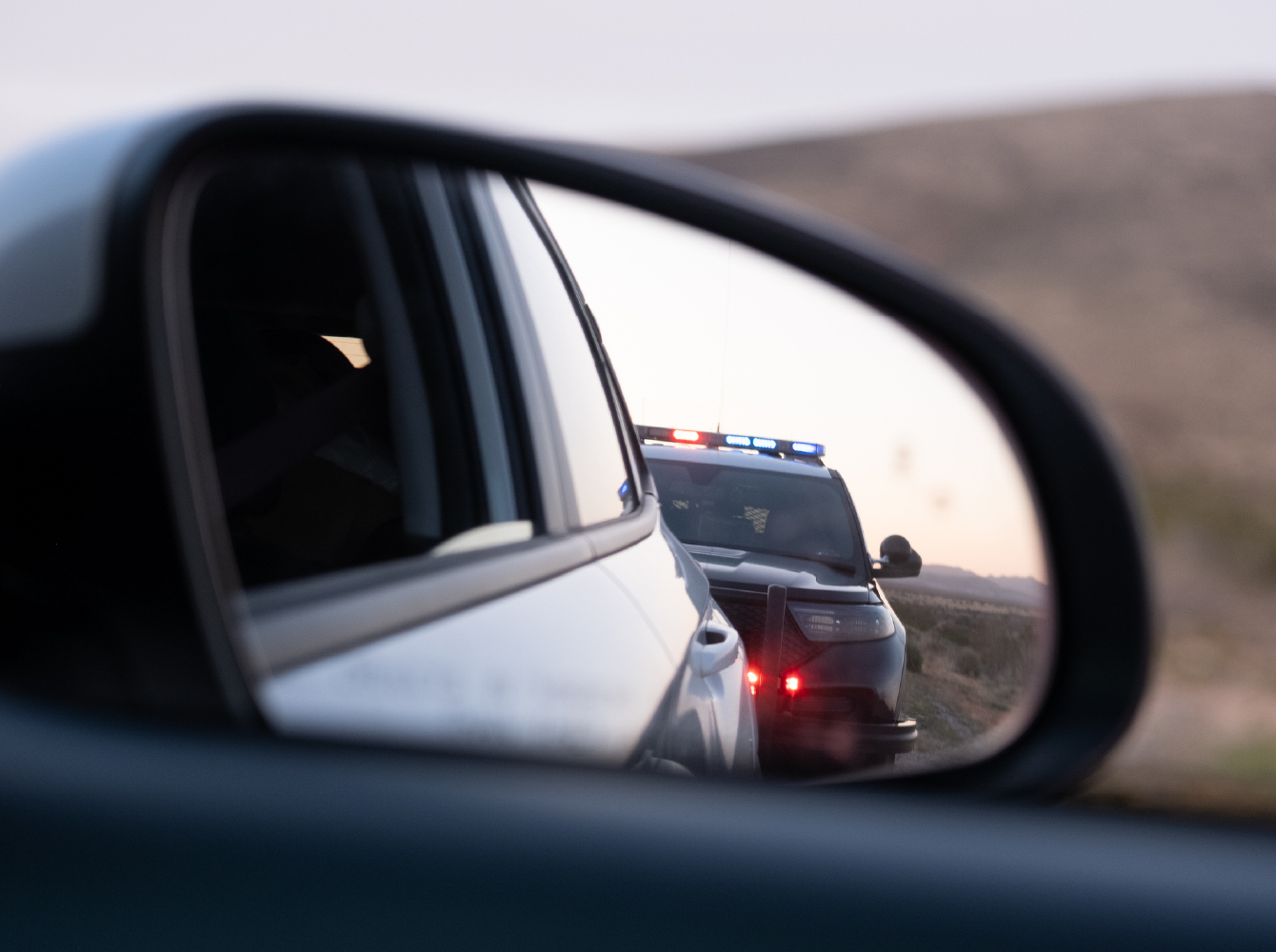 A police car with flashing lights reflected in a vehicle's side mirror on a rural road.