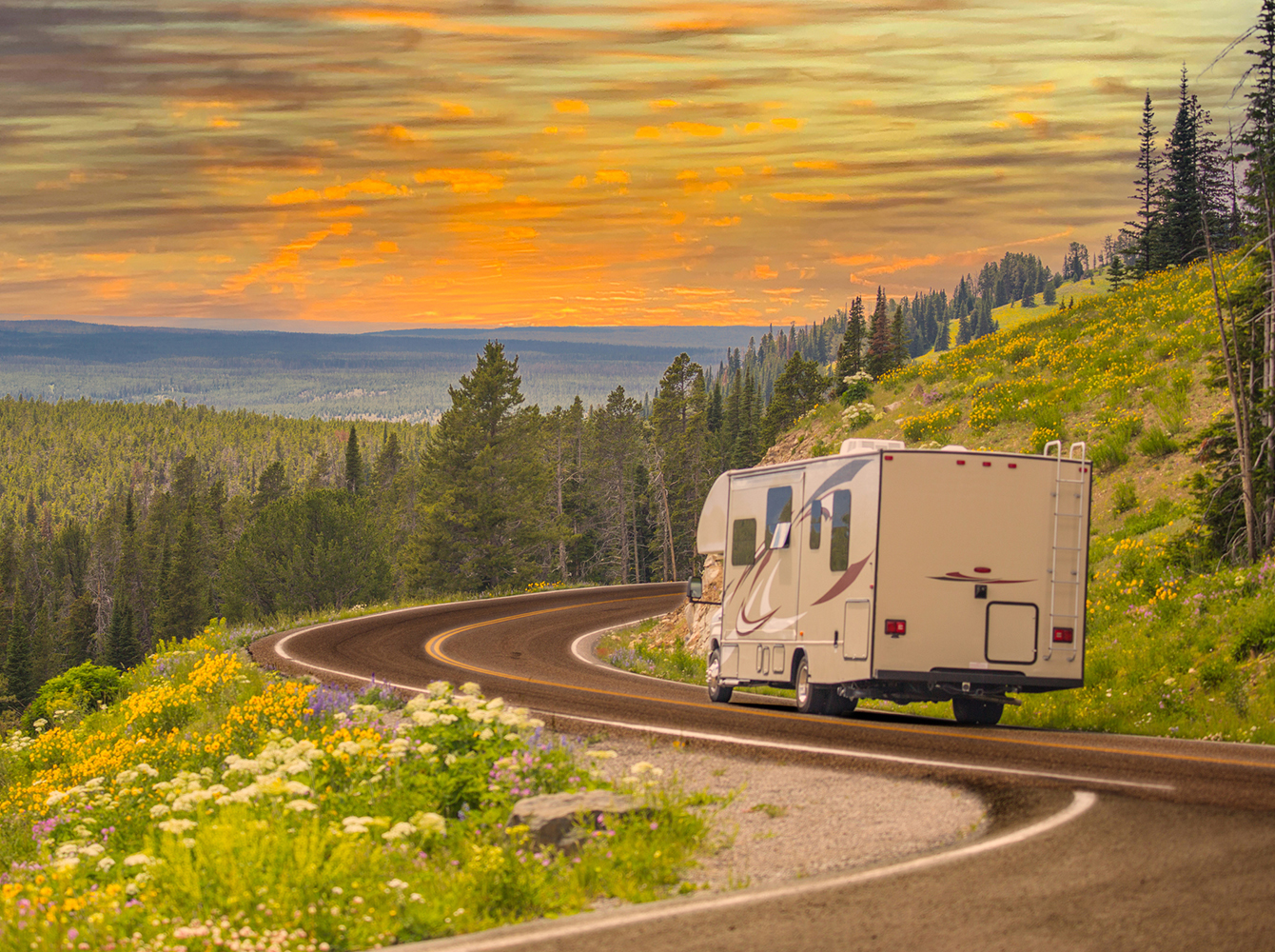 An RV driving along a winding mountain road lined with wildflowers and pine trees under a vibrant sunset sky.