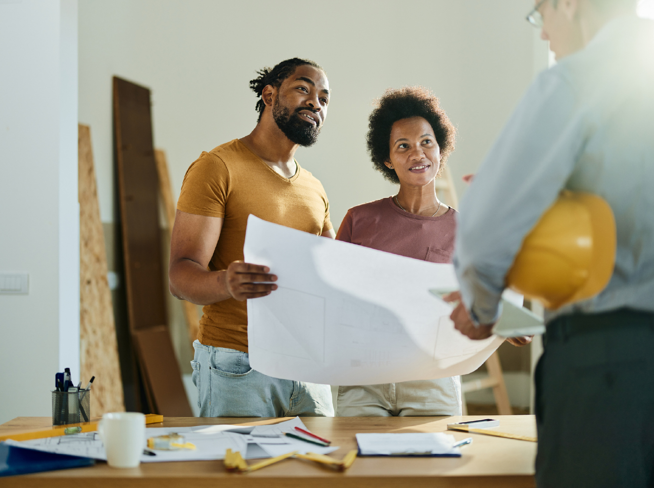 Two people reviewing blueprints with a contractor holding a hard hat in a home under renovation.