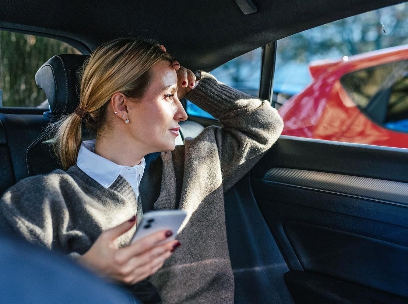 A woman is sitting in the back seat of a car holding her phone in her hand.