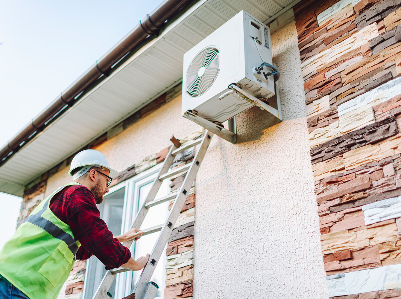 A worker climbs a ladder to reach an AC unit.