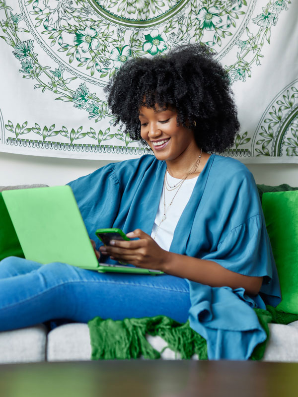 A woman sitting on a couch with her laptop on lap while using a phone