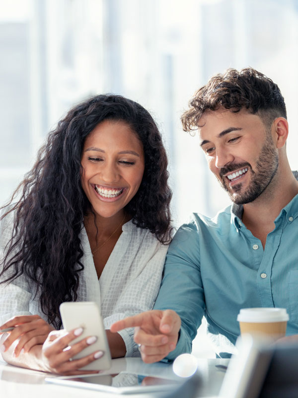 A man and a woman sitting at desk while staring and smiling at a phone