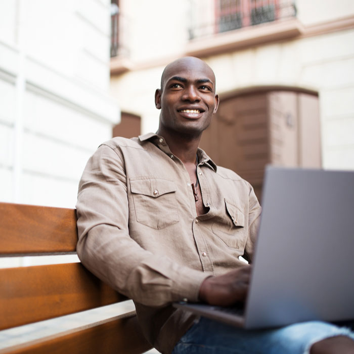 A smiling man using a laptop while sitting on a bench outside