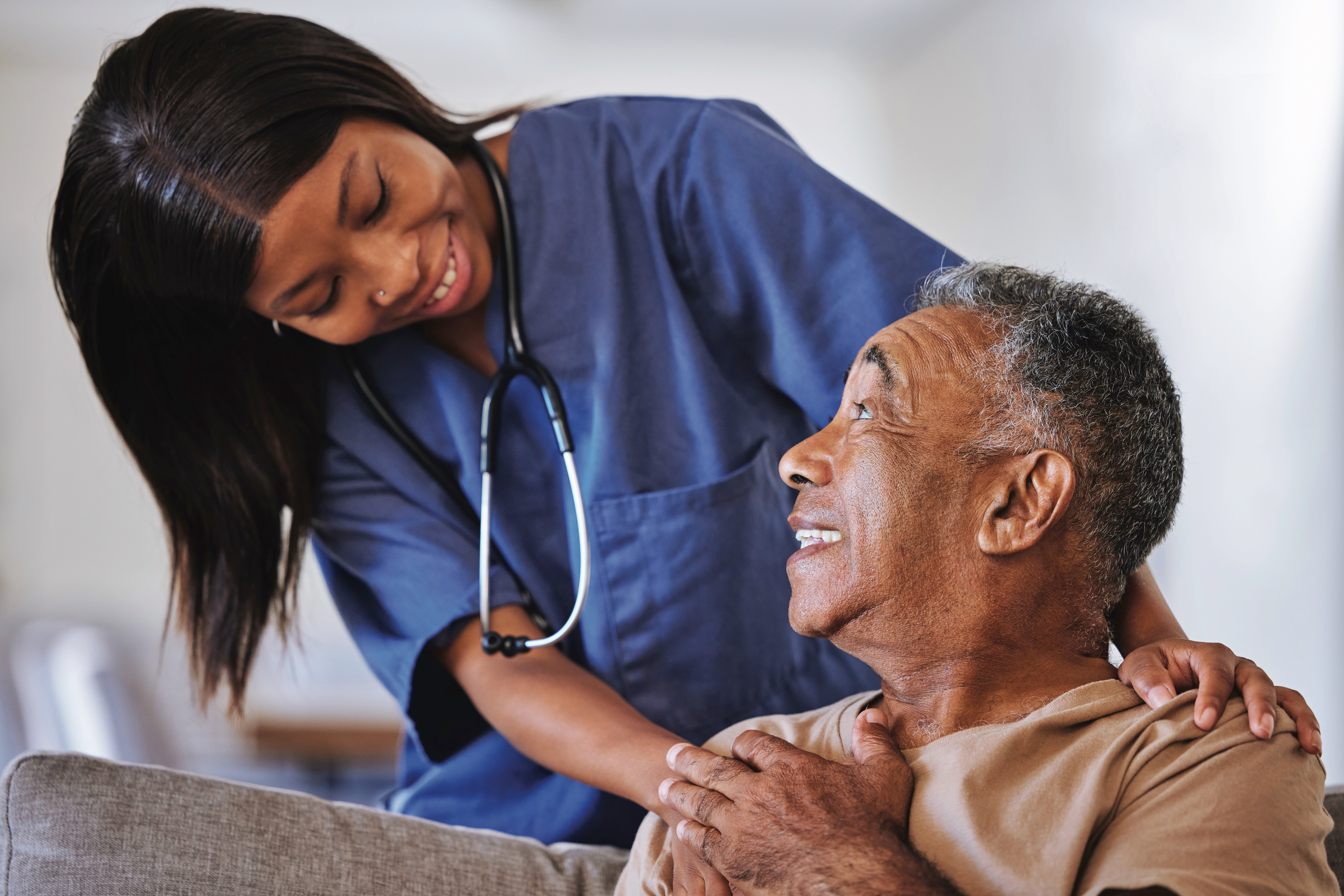 A female healthcare worker in blue scrubs smiles at and comforts an older adult male patient sitting on a couch