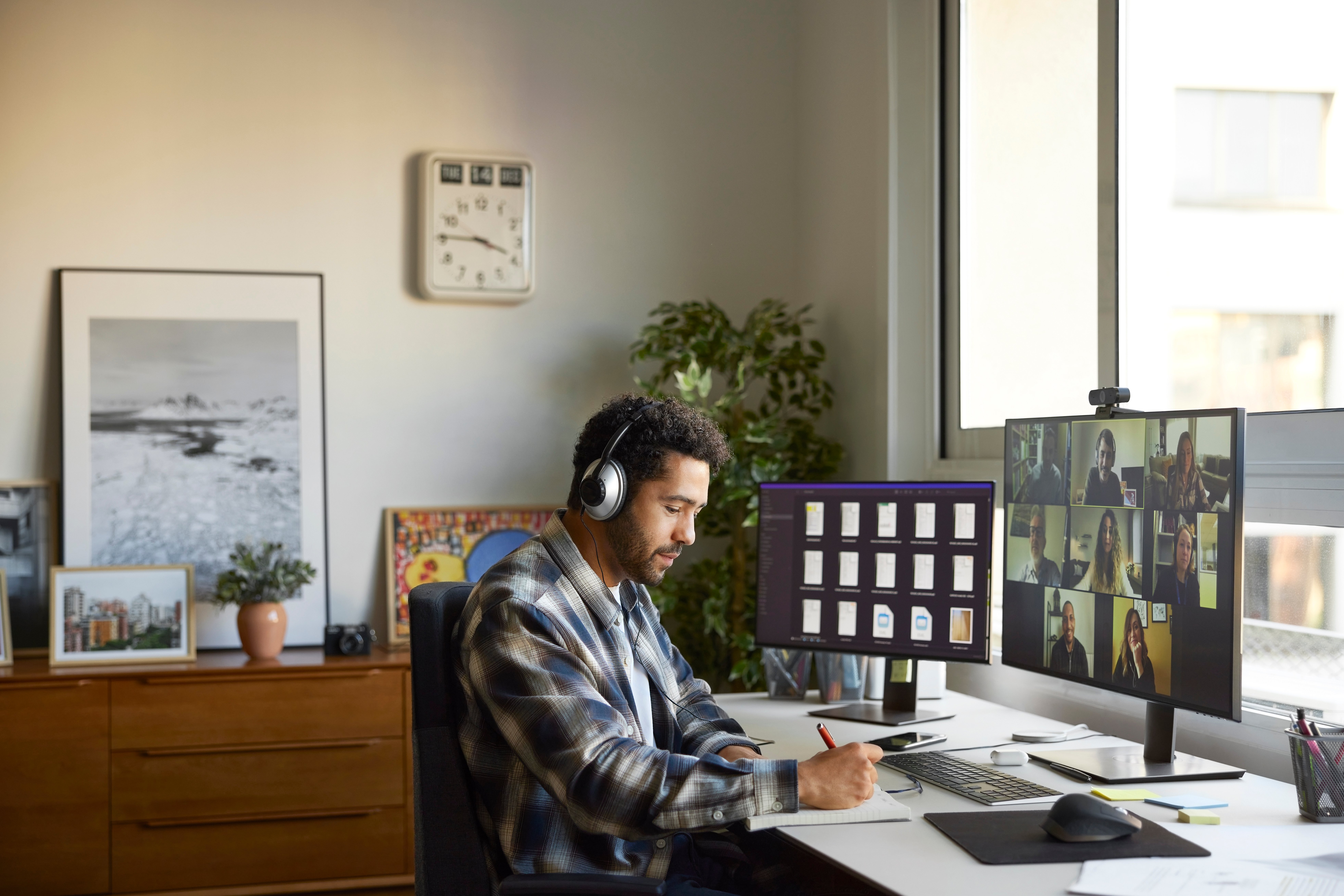 Man wearing headphones takes notes on paper while attending a virtual meeting.  
