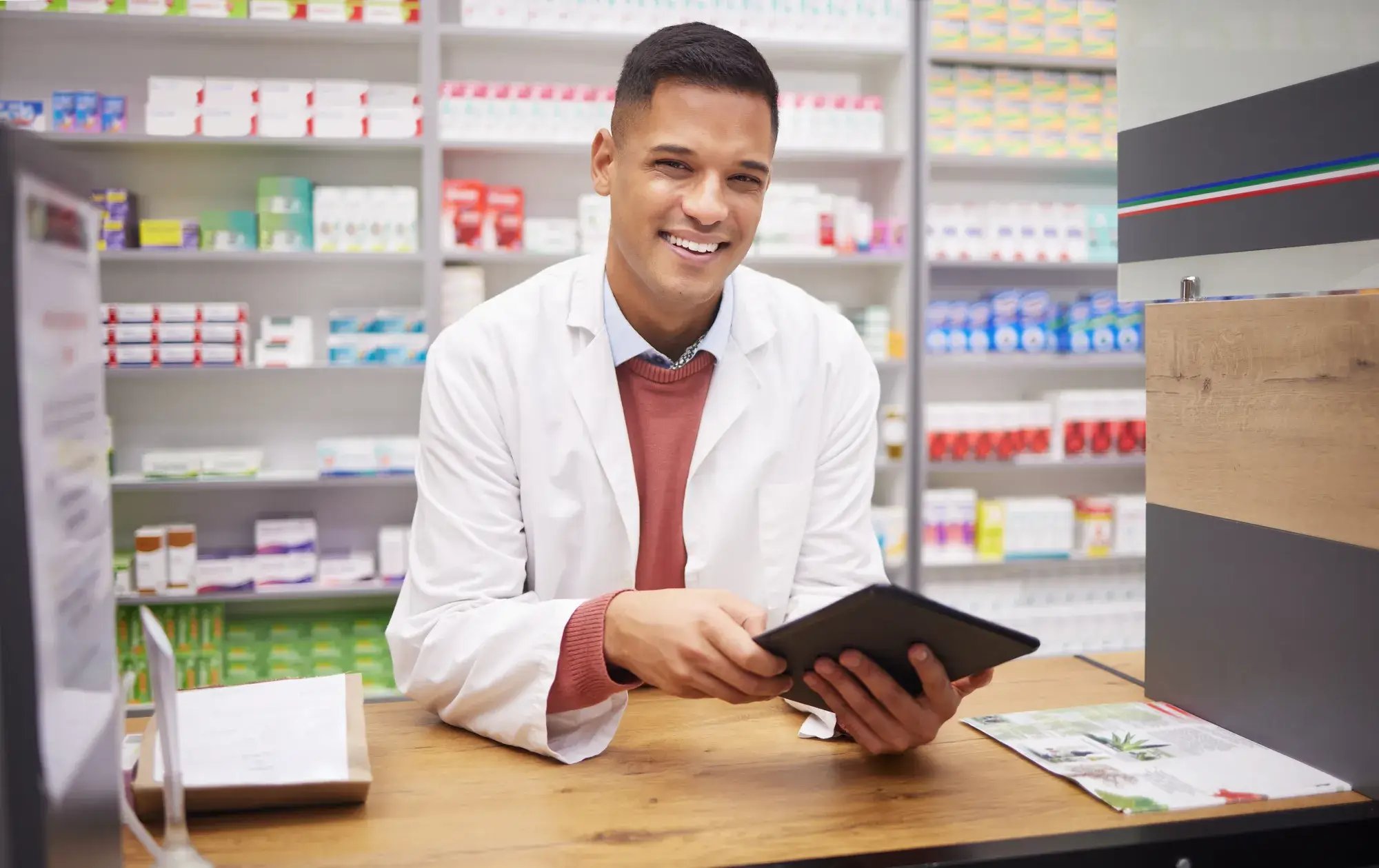 Smiling male pharmacist at counter with tablet in drugstore.