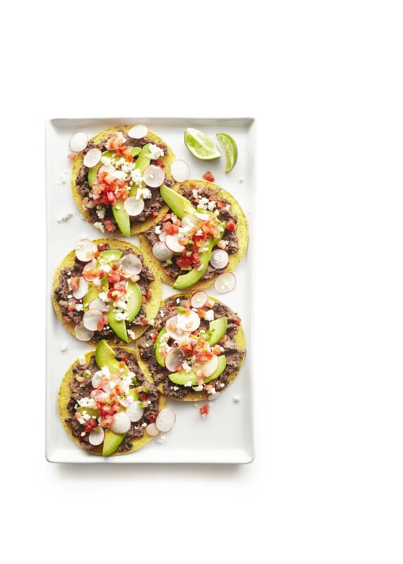 Overhead of a contemporary white rectangular platter with five tostadas with beans, radishes, coijta cheese, avacado, salsa, black beans and refried beans with limes on a white surface.
