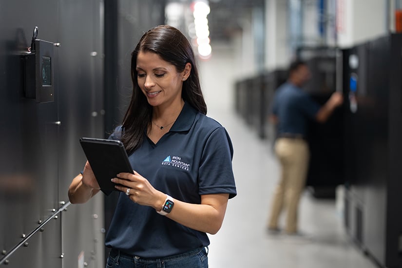 A female employee smiles while examining a device next to black cabinets in the data centre, reflecting Iron Mountain’s commitment to ensure secure erasure and provide data centre decommissioning services.