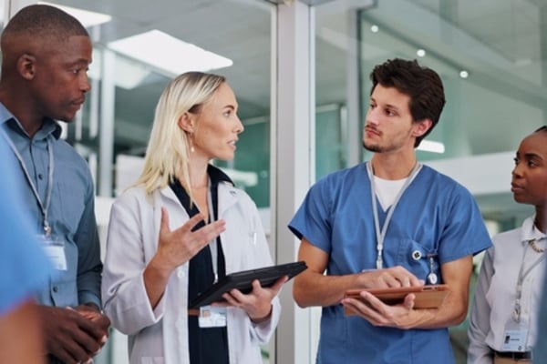 A diverse medical team in scrubs and lab coats gathers in a modern facility to discuss information on digital tablets.