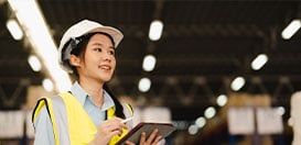 A female warehouse manager wearing a hard hat and safety vest while looking up with a digital tablet.